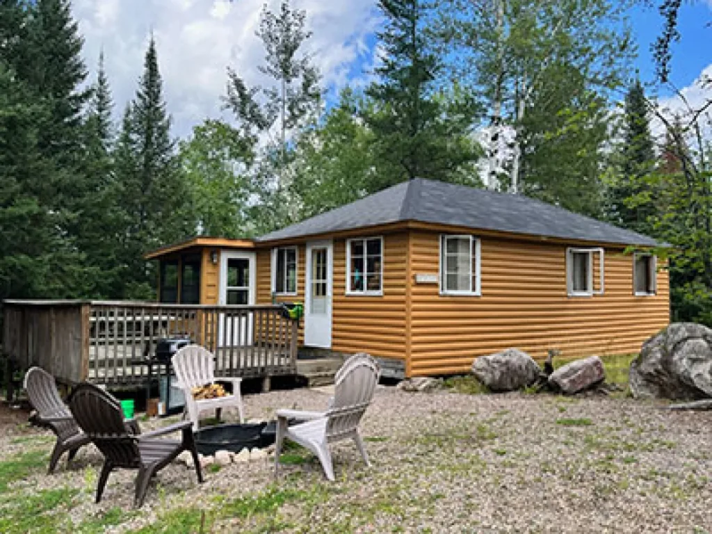 An outside view of the Alder cabin at Camp Northern Lights. It has a deck and lawn chairs around a fire pit.